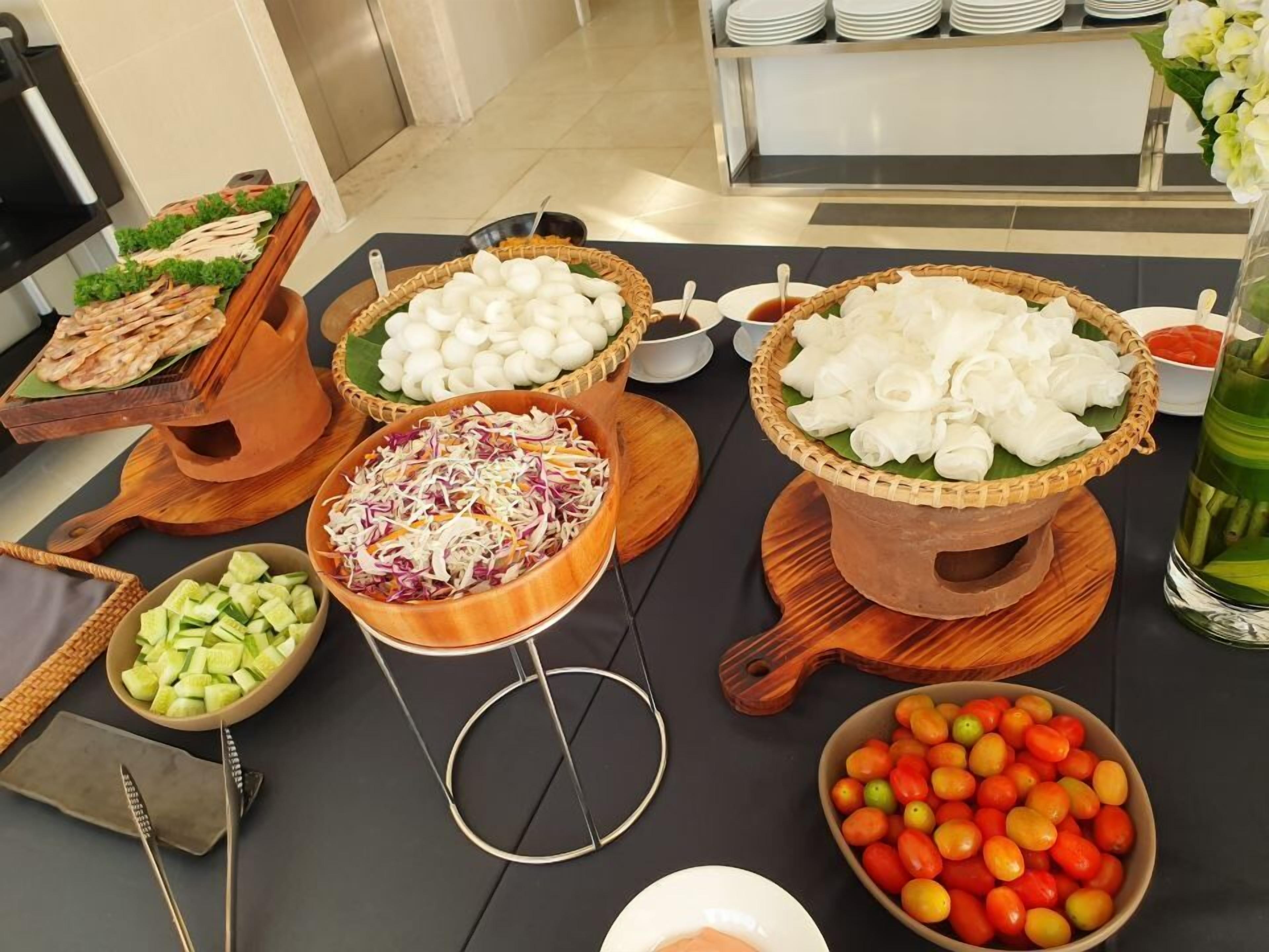 Una mesa de buffet con diversos alimentos, como ensalada de col, pepinos en rodajas, tomates cherry y albóndigas blancas sobre hojas de plátano.