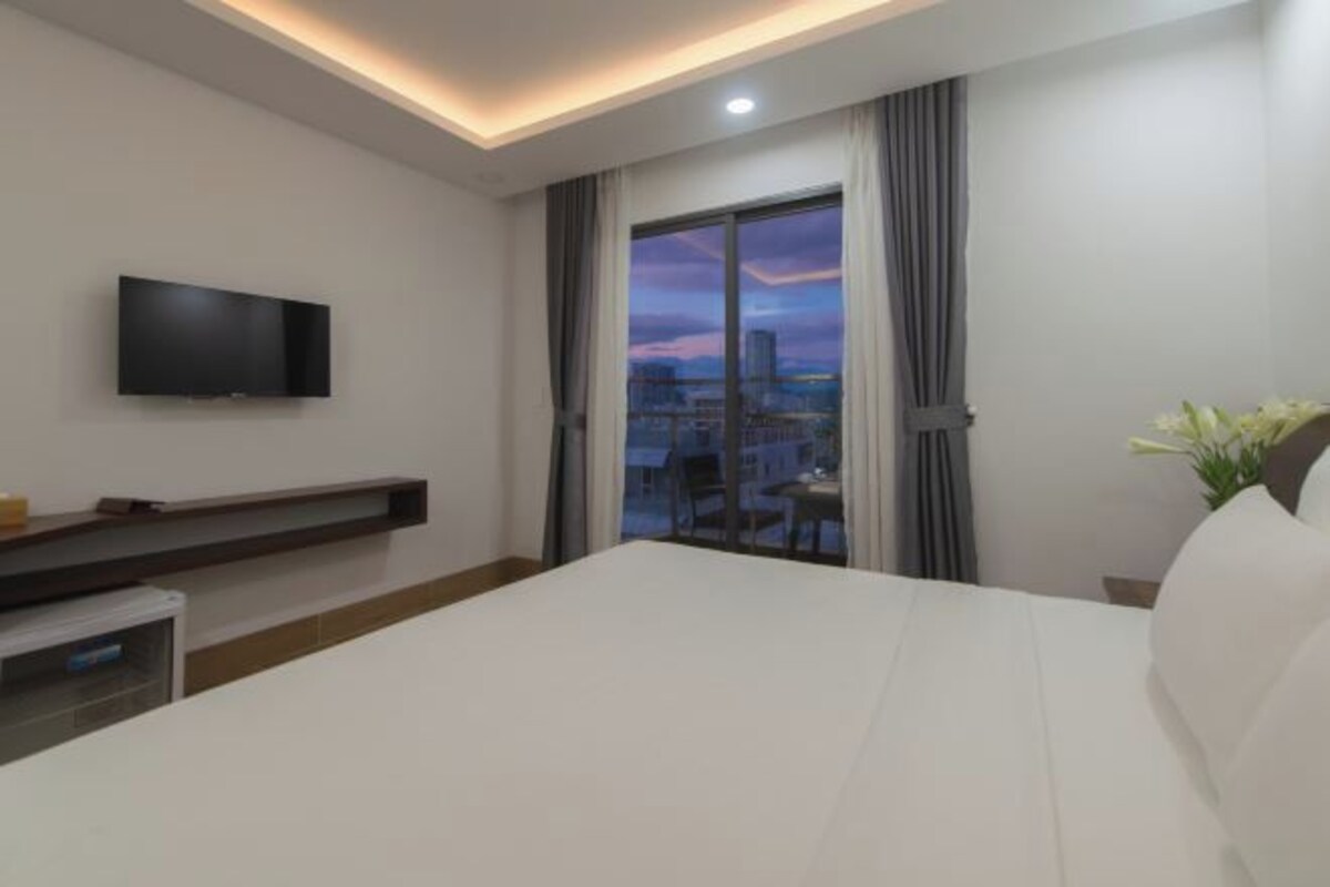 Una habitación de hotel moderna con una cama grande, un televisor de pantalla plana montado en la pared, una mesita pequeña y vistas a la ciudad a través de la ventana.