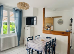 Een eetruimte met een tafel voor vier personen, een televisie op een kast en een raam met gordijnen.