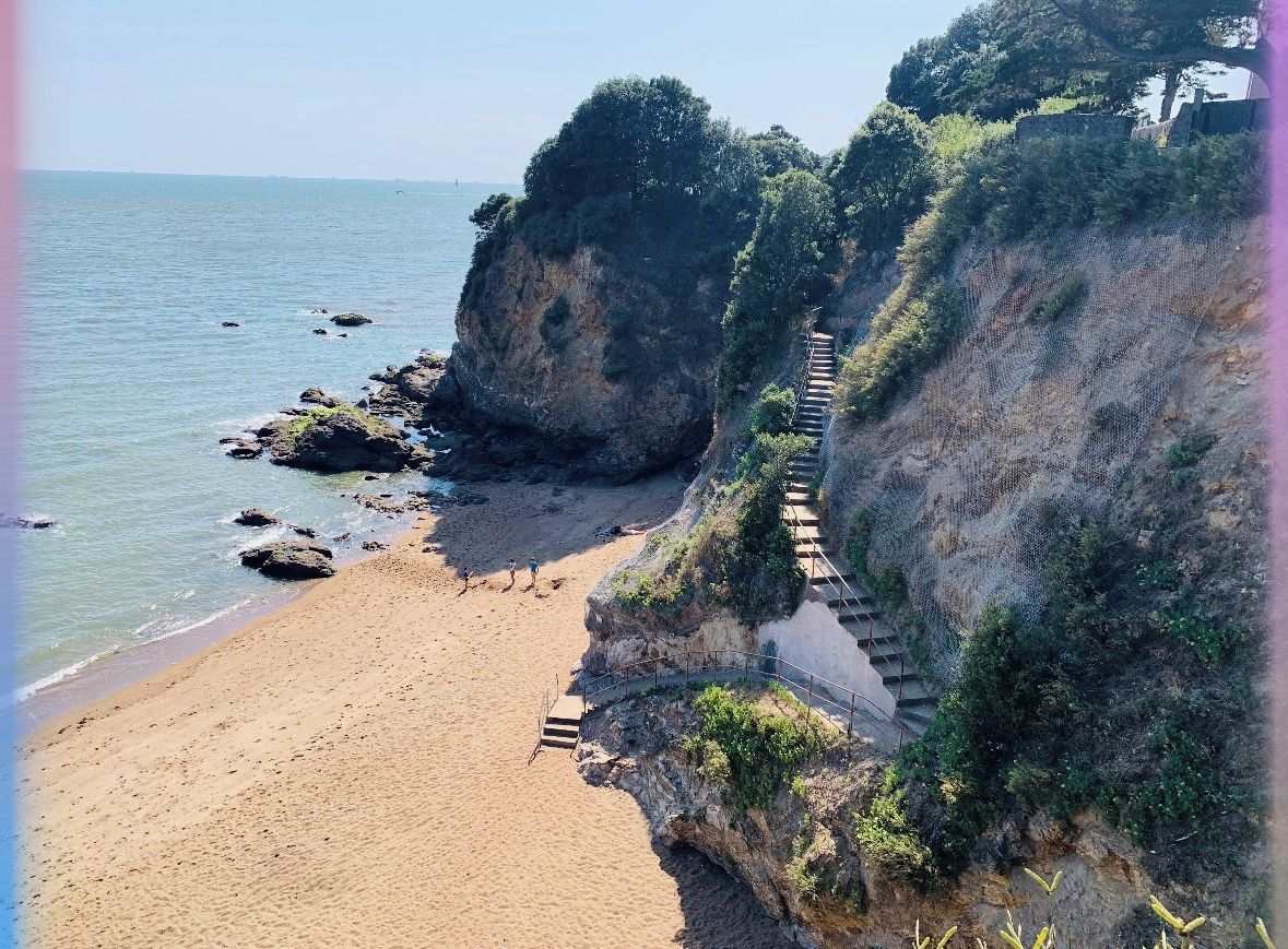 Une plage côtière avec un escalier descendant vers l’eau, entourée de falaises et de végétation.
