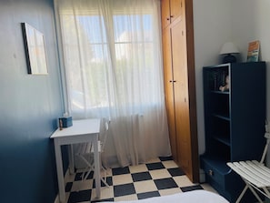 Ein Zimmer mit Schachbrettfußboden, einem Schreibtisch mit Stuhl, einem Kleiderschrank, einem Bücherregal und einem Fenster mit durchsichtigen Vorhängen.