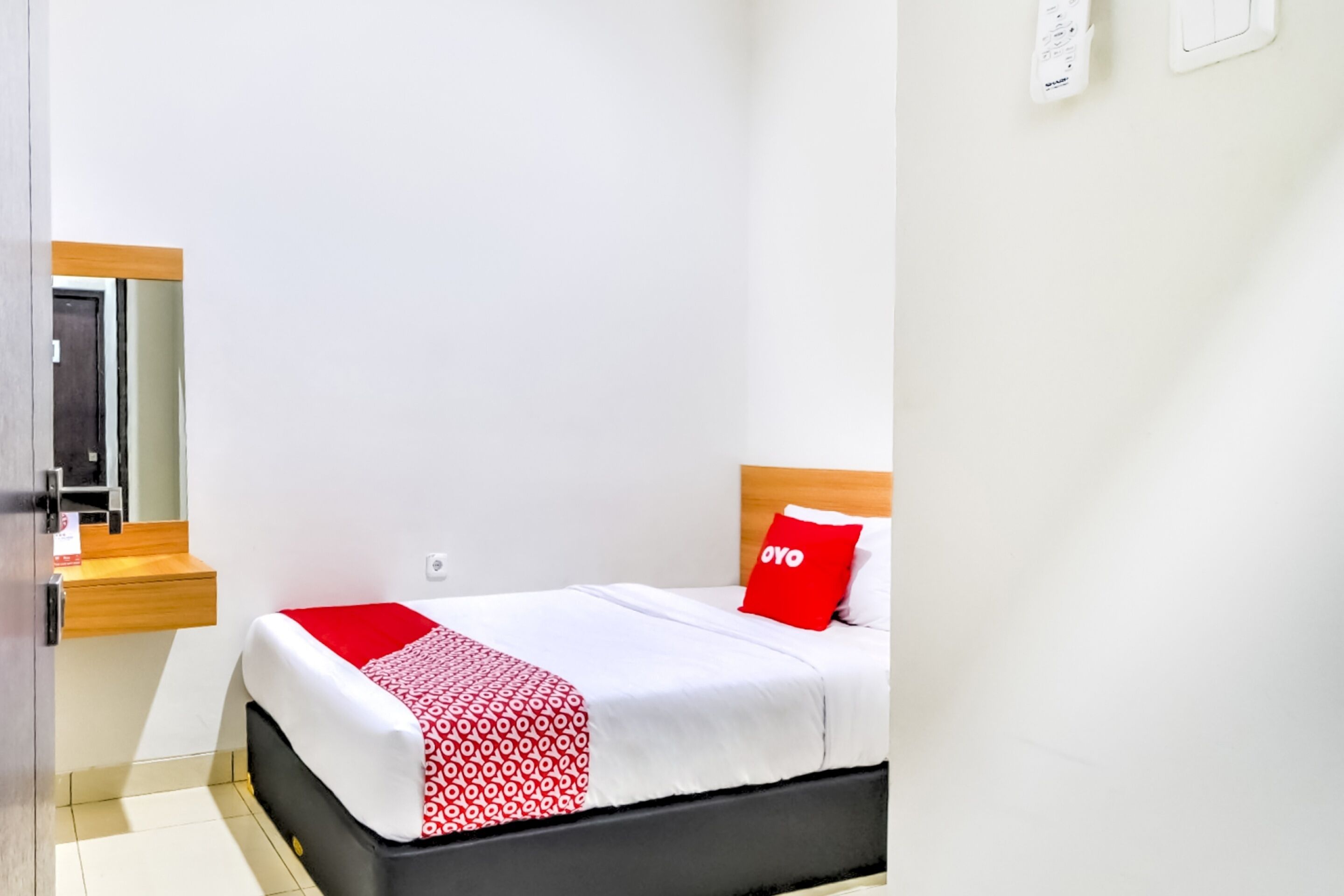 Quarto de hotel com uma cama, uma mesa de cabeceira de madeira, um espelho e um travesseiro vermelho.