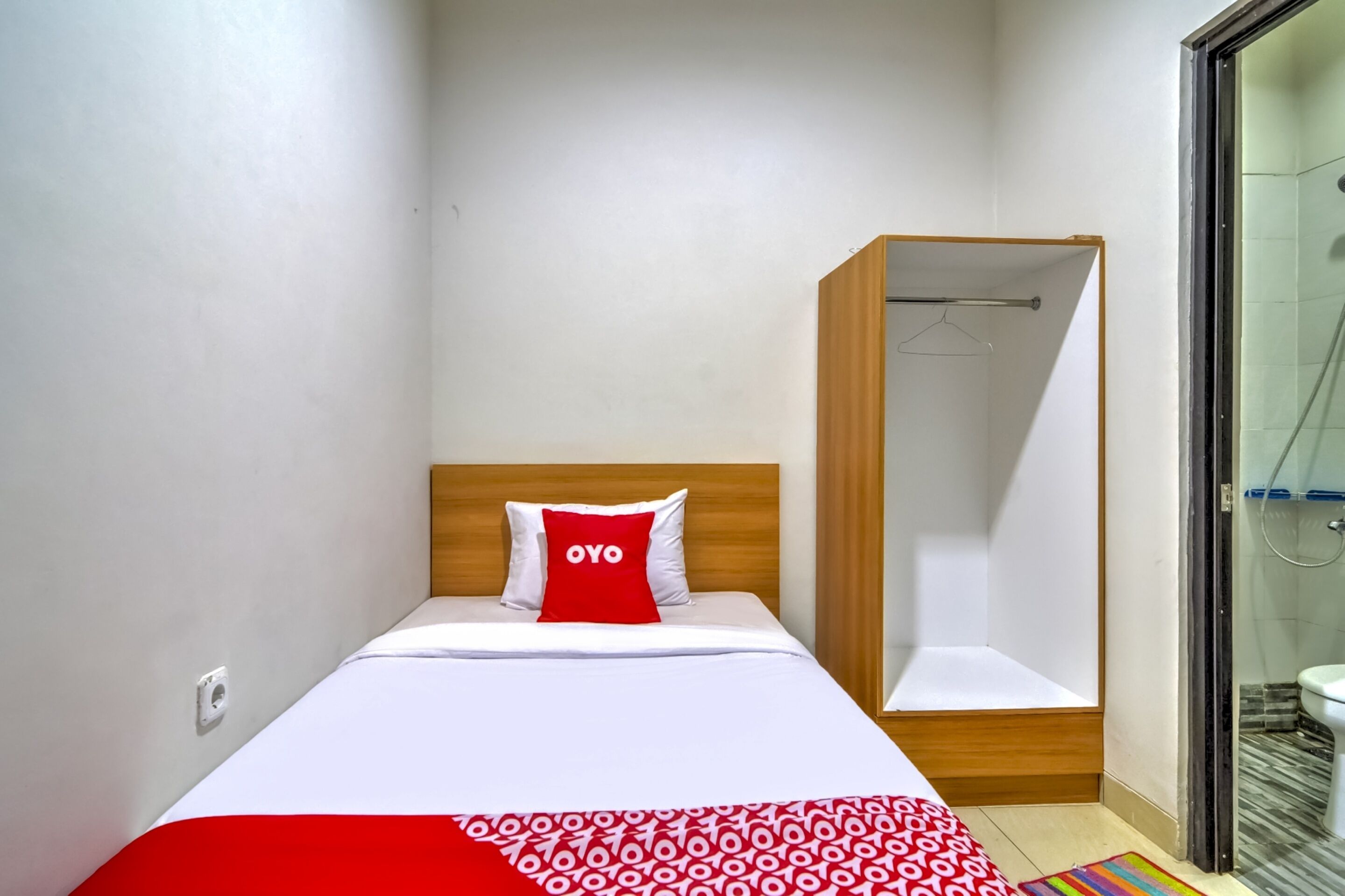A single bed with a red and white patterned bedspread, a wooden headboard, and a red pillow with the word "OYO" on it.