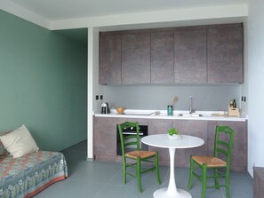 Una cocina compacta con encimera blanca, dos sillas verdes, una mesita redonda y una cama con colcha colorida.