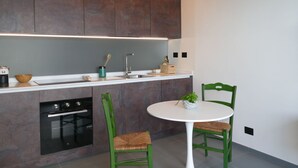 A modern kitchen with dark wooden cabinets, a white countertop, a built-in oven, and a round table with two green chairs. - Beautiful apartment for 3 guests with WIFI and terrace (Ventimiglia)