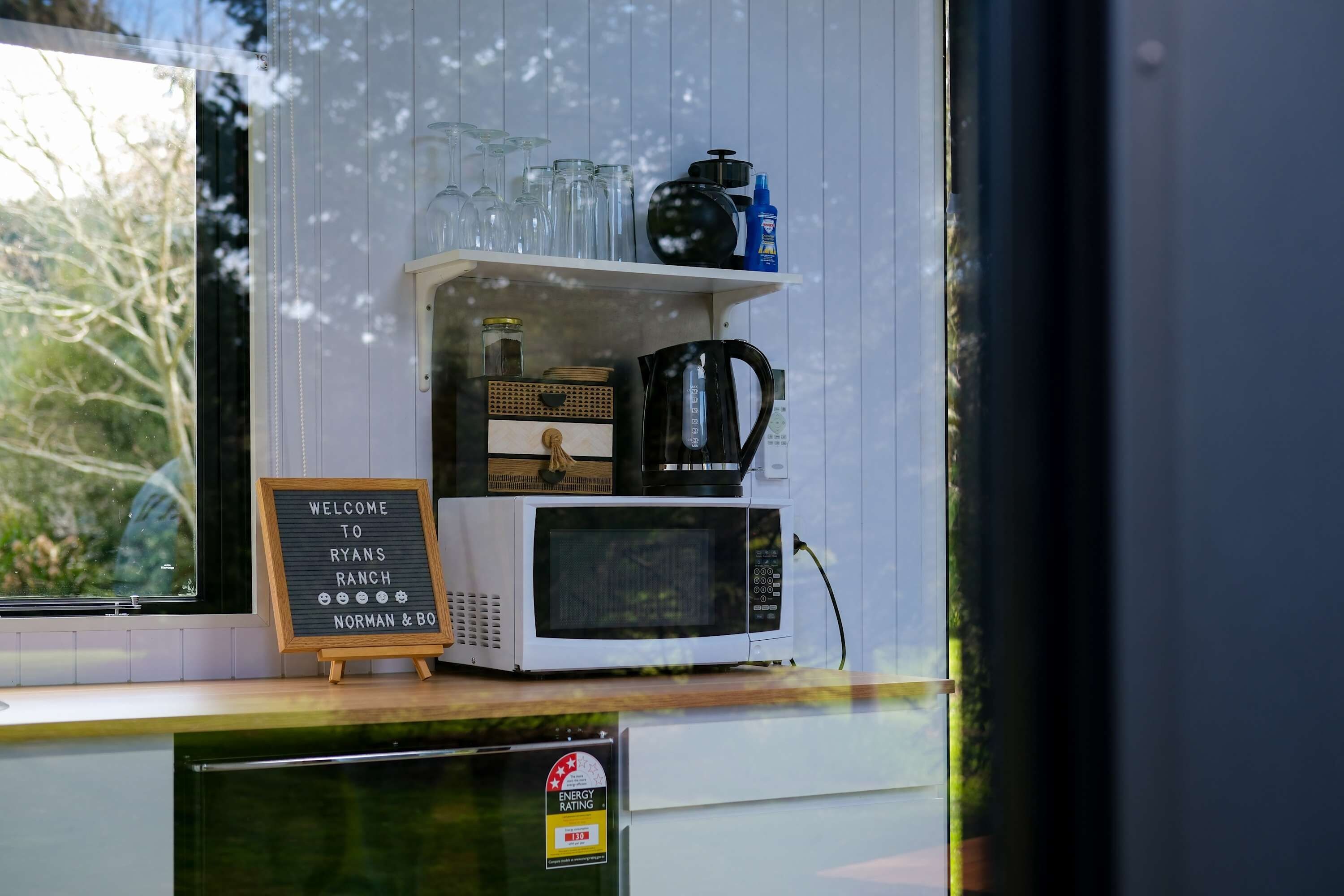 Una encimera de cocina con un microondas blanco, una cafetera y una estantería con cristalería y una botella azul. Un letrero dice 'Welcome to Ryan's Ranch Norman & Boone'.