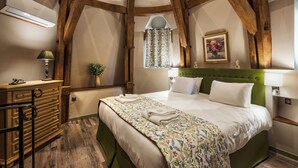 Un dormitorio con techo de madera, una cama con cabecera verde, una mesita de noche con lámpara, un aparador y una ventana con cortinas estampadas.