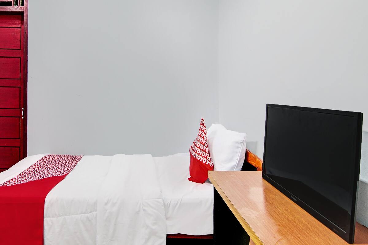 A single bed with a white and red bedspread, a red pillow, and a black television on a wooden table beside the bed.