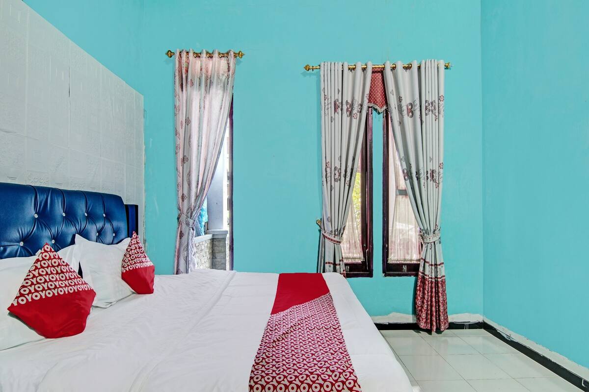 A bedroom with a blue headboard, white bedding, red pillows, and patterned curtains.
