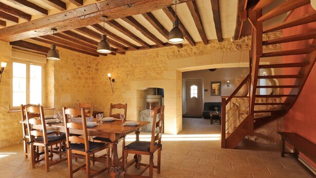 A rustic dining area with a wooden table and chairs, a stone wall, and a wooden staircase.