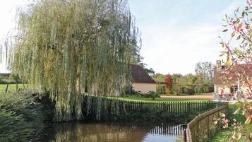 A serene riverside scene with a weeping willow tree, a small house with a garden, and a wooden fence.
