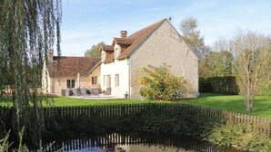 A stone house with a pond in front, surrounded by greenery and a wooden fence.