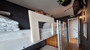 Ein Schlafzimmer mit Etagenbett, einer Holzleiter und einem weißen Kopfteil. Eine Tür führt in einen weiteren Raum, und an der Wand hängt ein Spiegel.