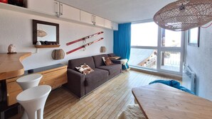 Ein modernes Wohnzimmer mit Holzboden, einem braunen Sofa, weißen Beistelltischen und einem großen Fenster mit Blick auf eine Holzterrasse.