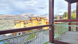 Ein Holzbalkon mit Metallgeländer, der auf eine gebirgige Landschaft und ein Gebäude mit vielen Fenstern blickt.