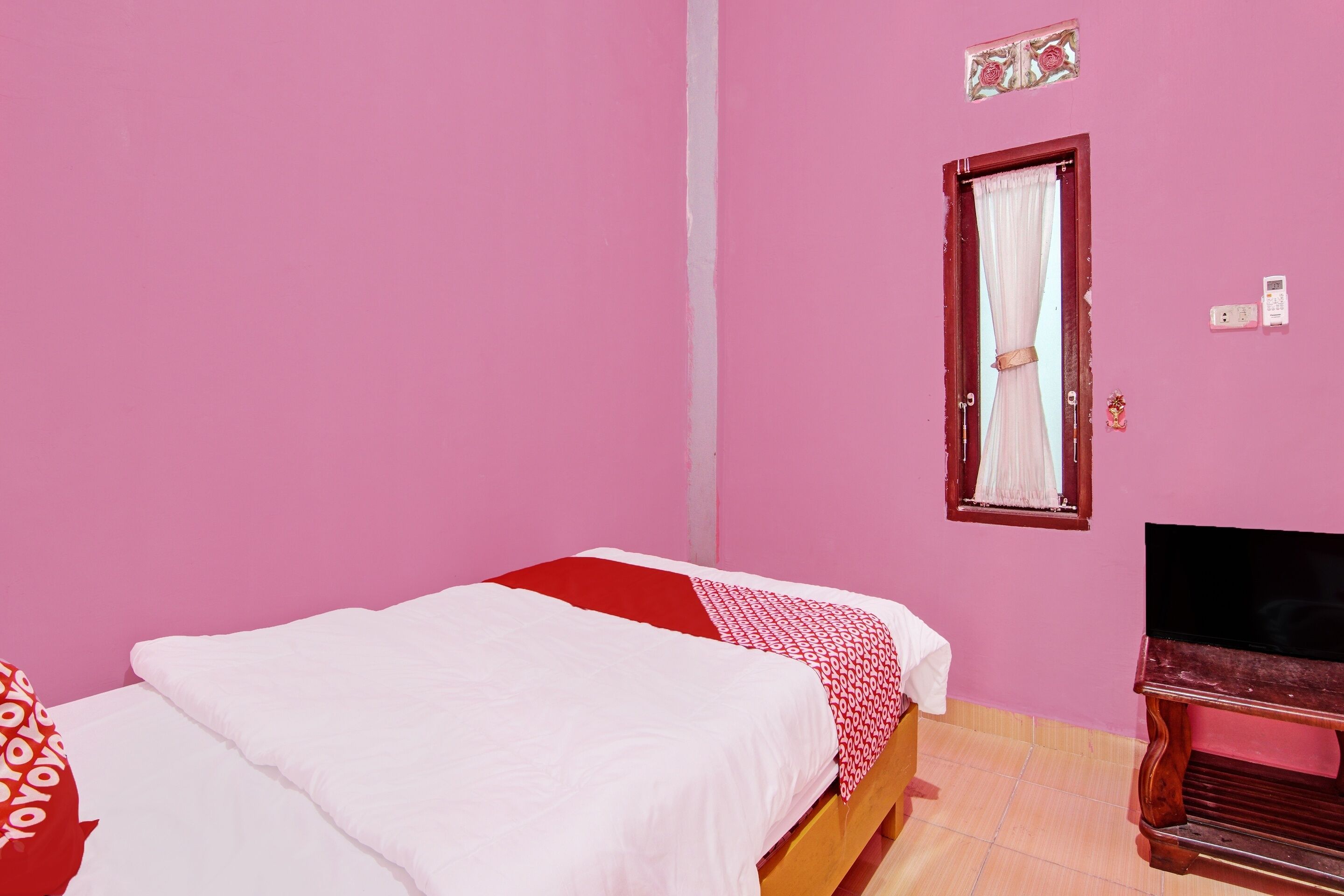 Una habitación con paredes rosas, una cama con ropa de cama blanca y roja, un banco de madera, una ventana pequeña con cortina y un enchufe de pared.
