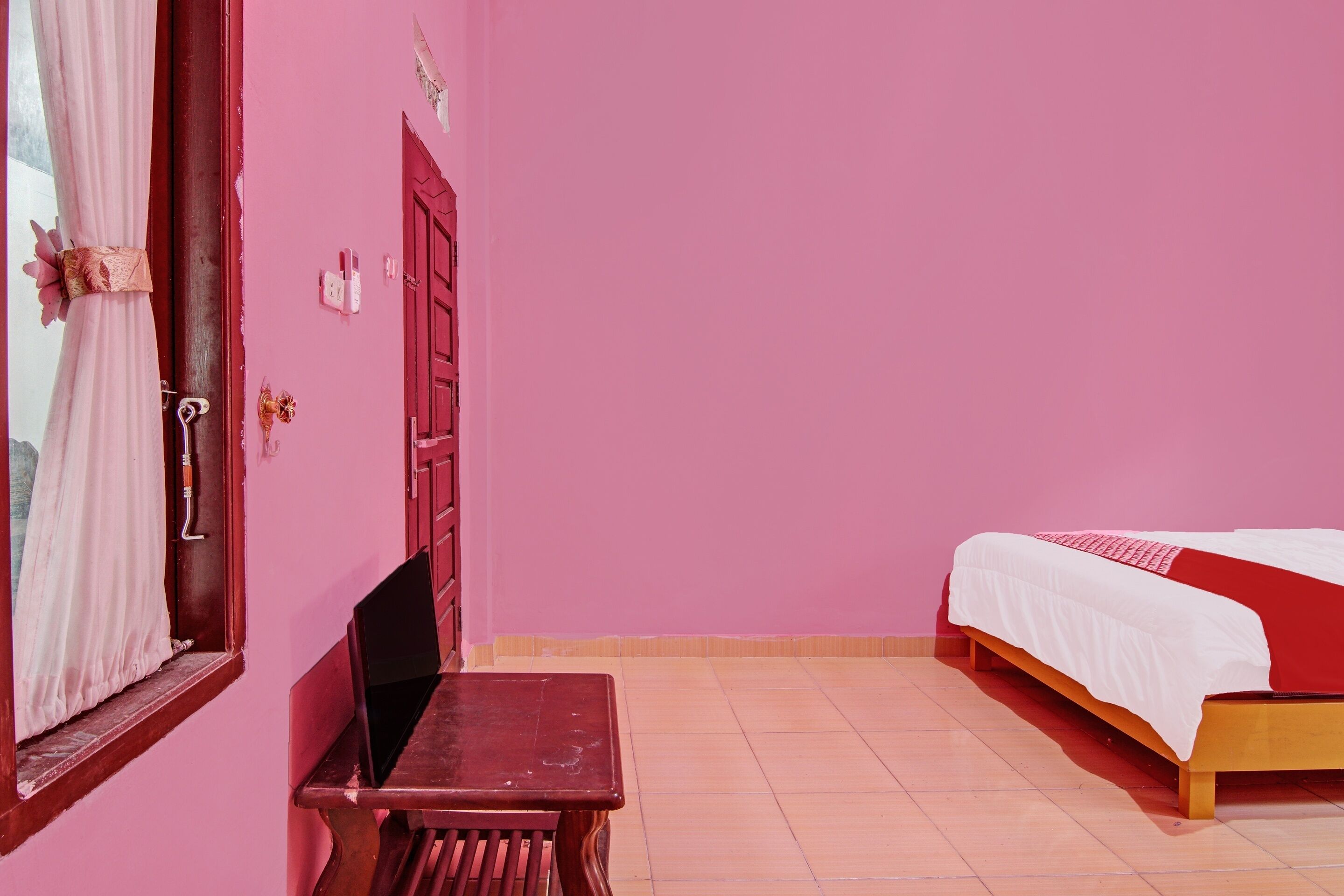 A room with a bed, a wooden table, a window with curtains, and a red door.
