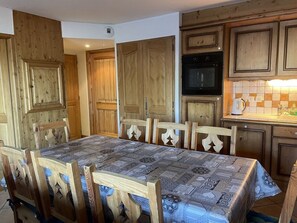 Un comedor con una mesa cubierta por un mantel estampado, sillas de madera y una cocina con armarios de madera y un microondas.