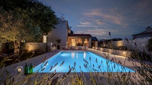 Zona de piscina amb mur de pedra, casa amb teulada enrajolada i mobiliari exterior.