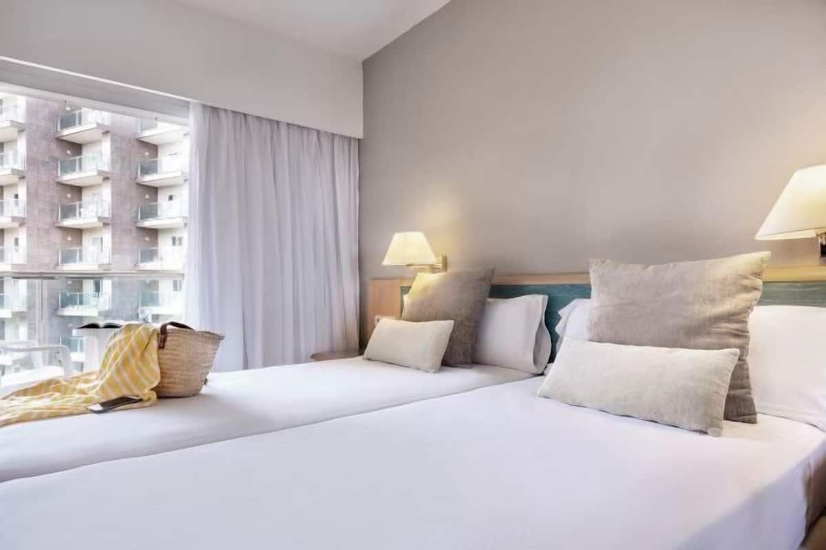 A hotel room with two beds, a window with curtains, a lamp on each side, and a view of a building outside.