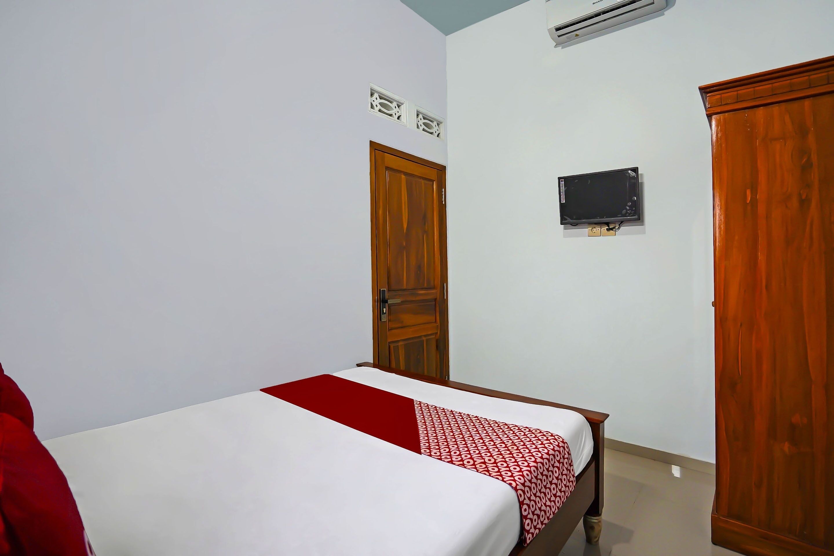 A bedroom with a bed, a wooden wardrobe, a television on the wall, and an air conditioning unit.