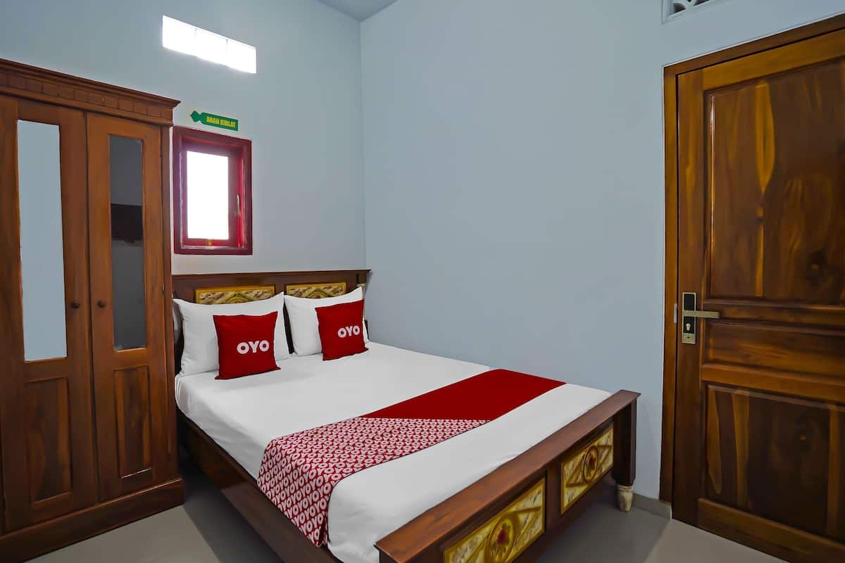 Una camera da letto con un letto in legno, due cuscini rossi, una coperta bianca e una coperta rossa. Si nota un armadio in legno con uno specchio e una piccola finestra con cornice rossa.