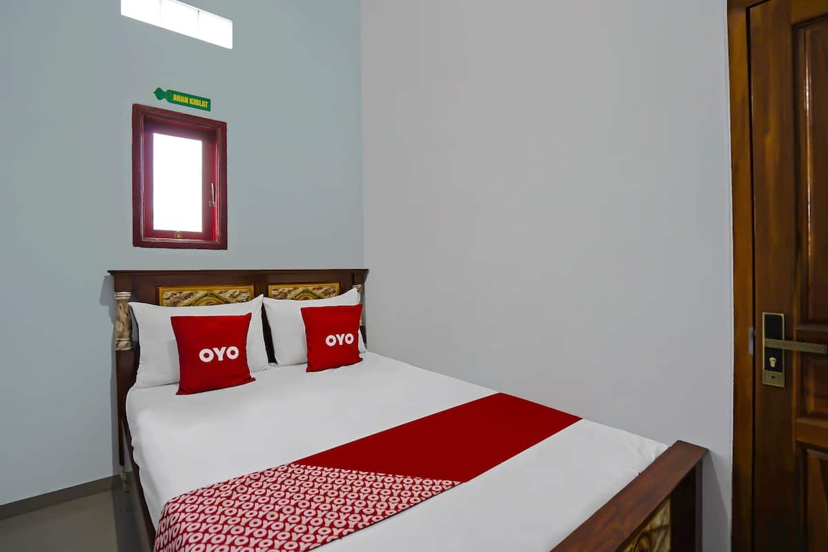 Una camera d'albergo con un letto, due cuscini rossi, una coperta a motivi rossi e bianchi, una testiera in legno, una finestra con vista e una porta.