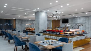 A modern dining area with multiple tables and chairs, a buffet with various food items, and a contemporary ceiling design.