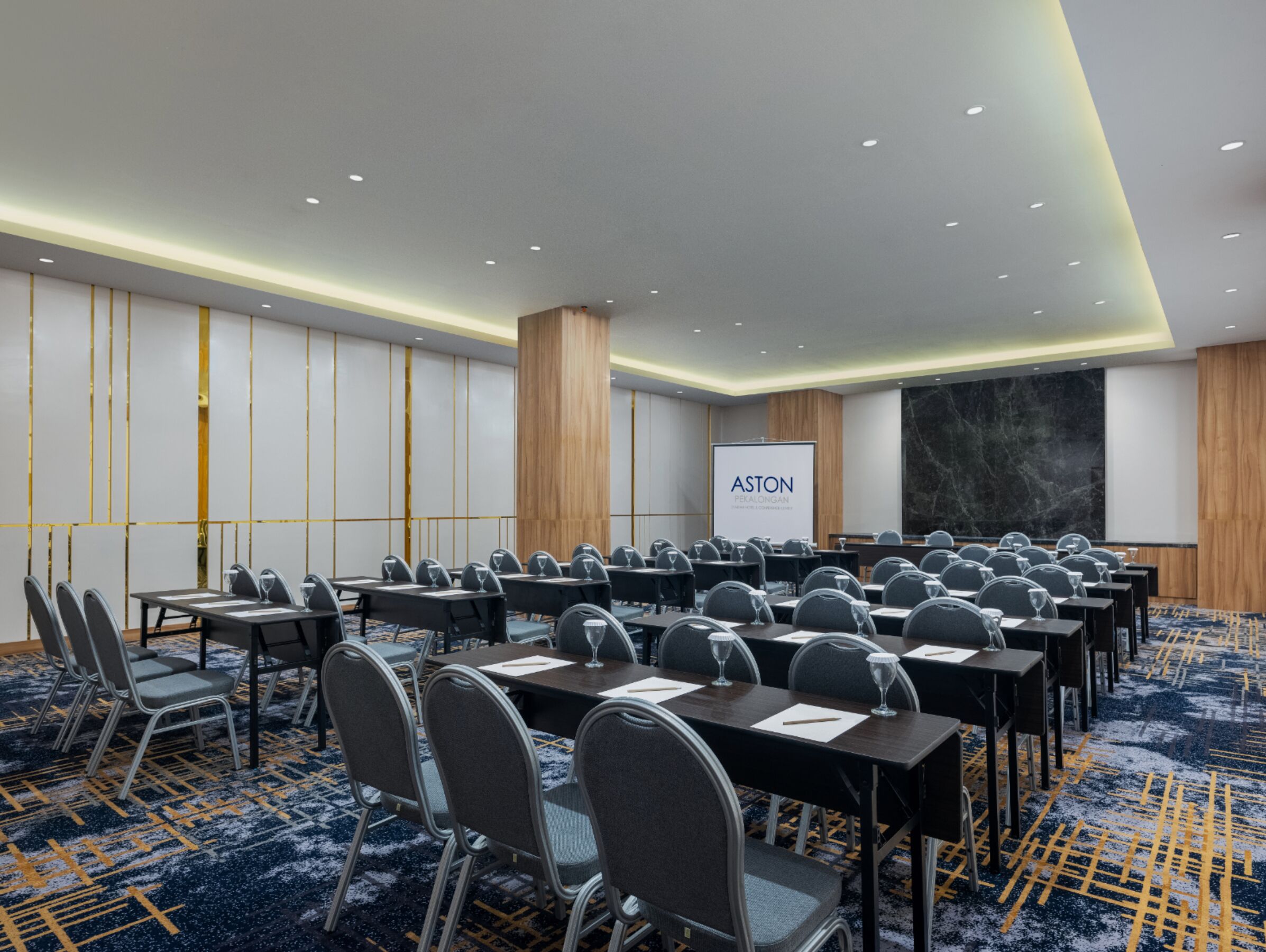 A conference room with rows of tables and chairs, a large screen, and a marble wall.