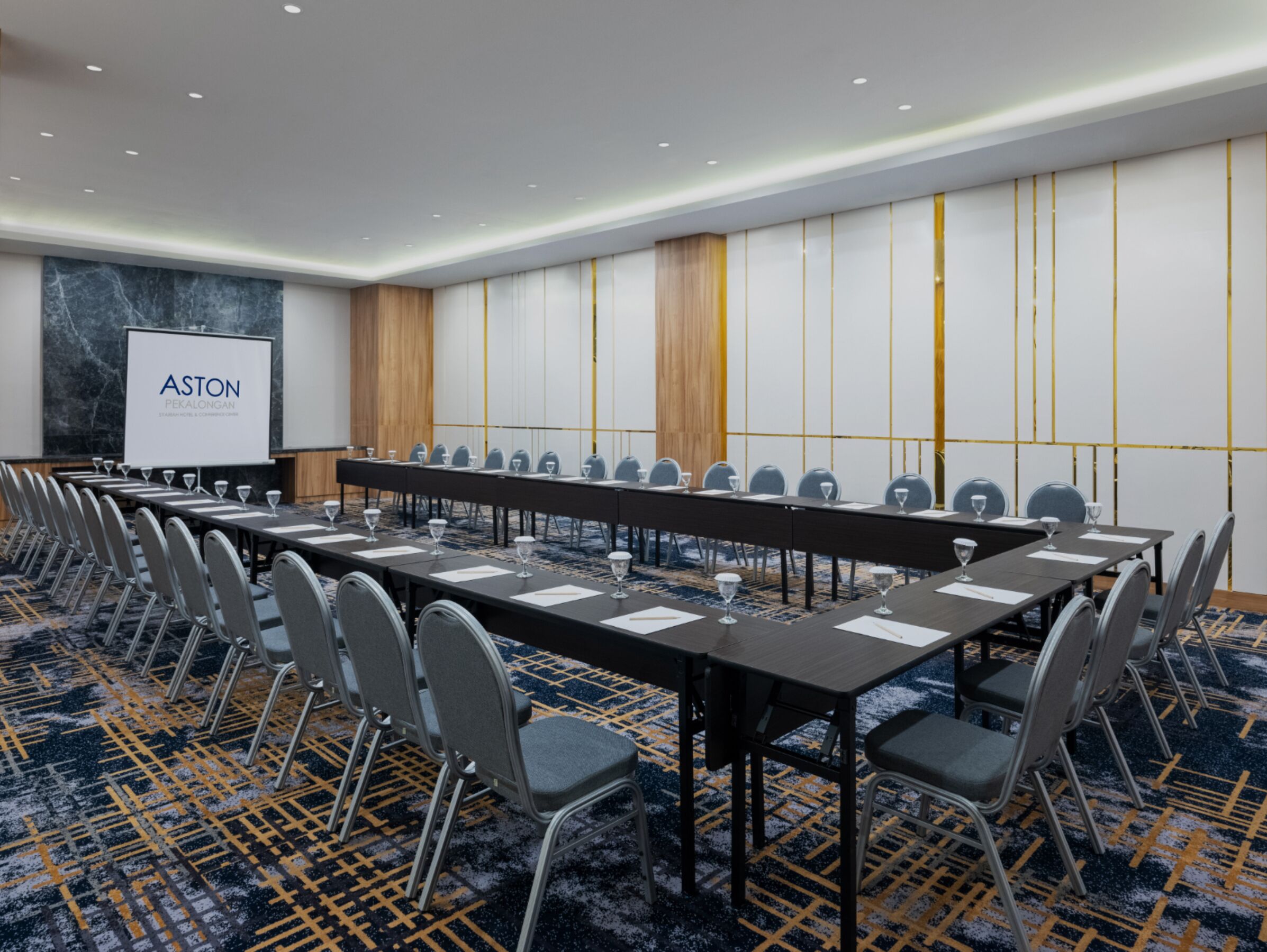 A conference room with a long table, chairs, and a projector screen displaying "ASTON PEKALONGAN".