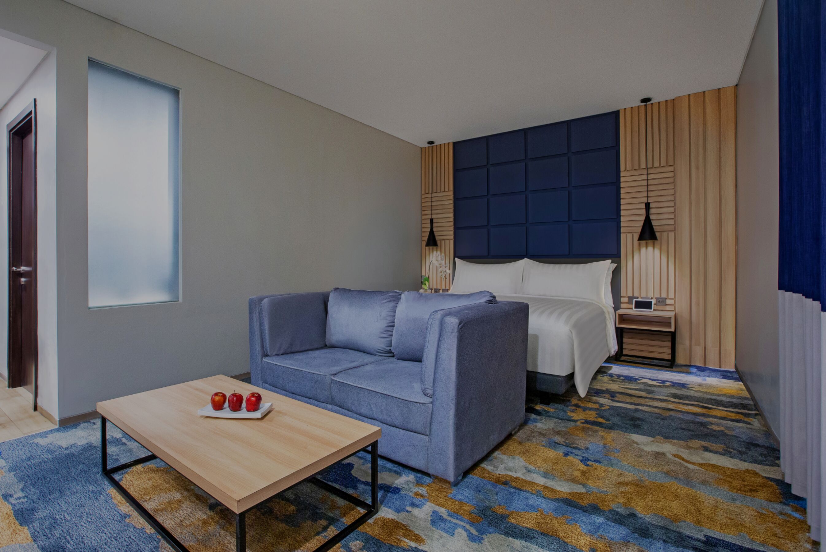 A modern hotel room with a blue sofa, a wooden coffee table, a bed with a blue headboard, and a patterned carpet.