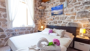 Ein Schlafzimmer mit Steinwand, einem Bett mit weißen Leinentextilien, einem Kopfteil, zwei Nachttischen mit Lampen und einem Fenster mit Vorhängen.