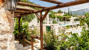 Ein Steinhaus mit einer Holz-Pergola, einer Steintreppe und Blick auf Pool und Berge im Hintergrund.