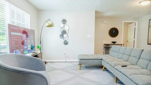 A modern living room with a sectional sofa, a grey armchair, a patterned rug, a wall-mounted lamp, and a decorative wall piece.