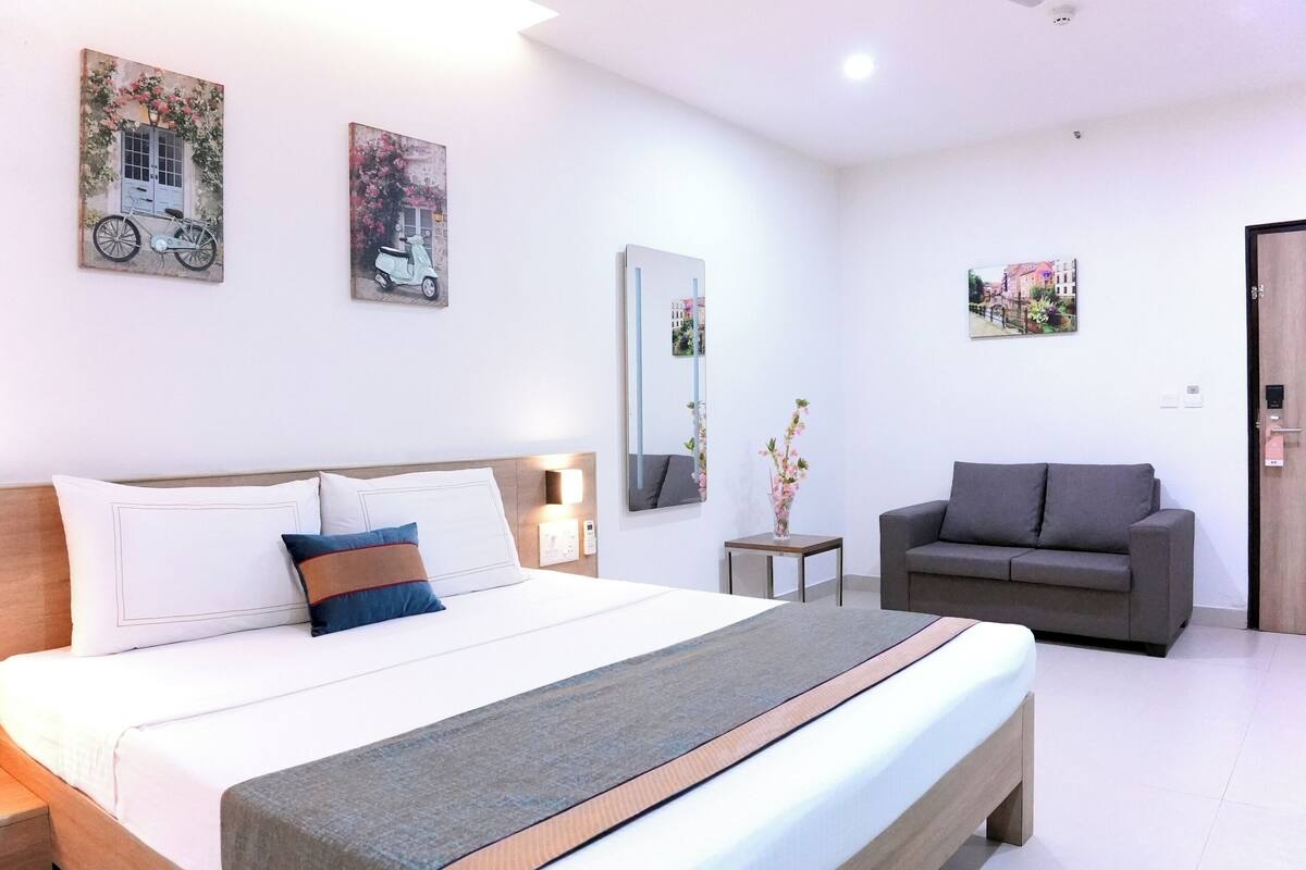 A hotel room with a bed, a gray sofa, a small table with a vase, and artwork on the walls.