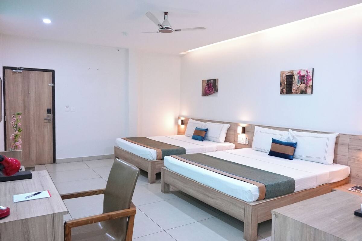 A hotel room with two beds, a desk, a chair, a ceiling fan, and wall-mounted lights.