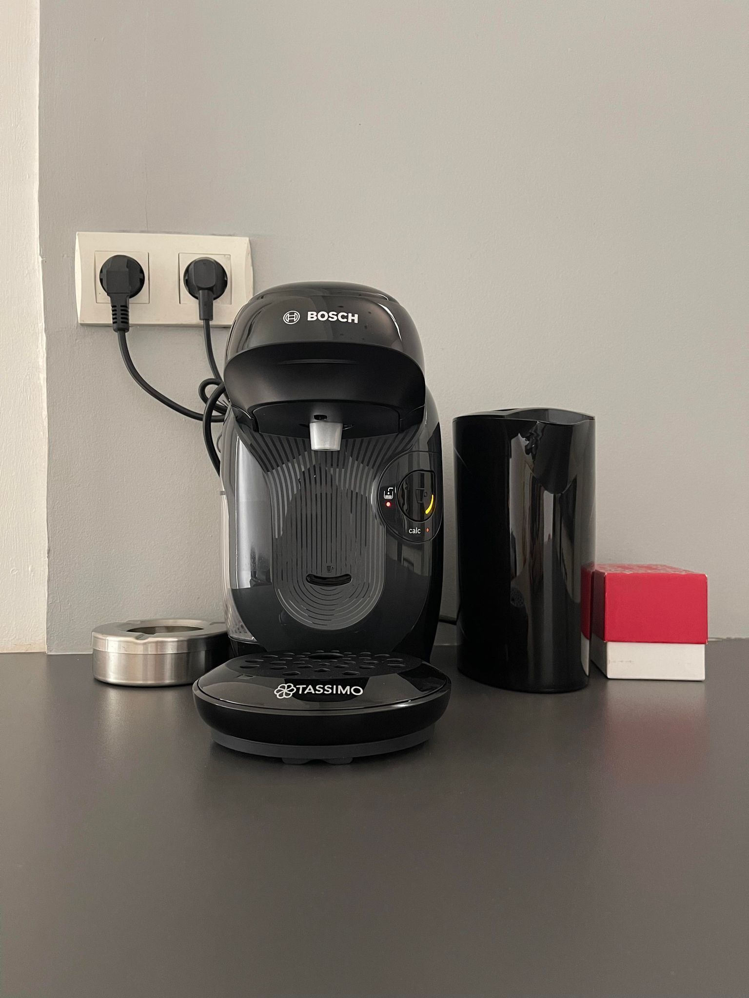 Une machine à café avec un réservoir d’eau, un câble d’alimentation et une boîte rouge, le tout posé sur une table.