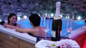 A bottle of wine and two glasses on a table with a flower arrangement, in front of a hot tub with people in the background.