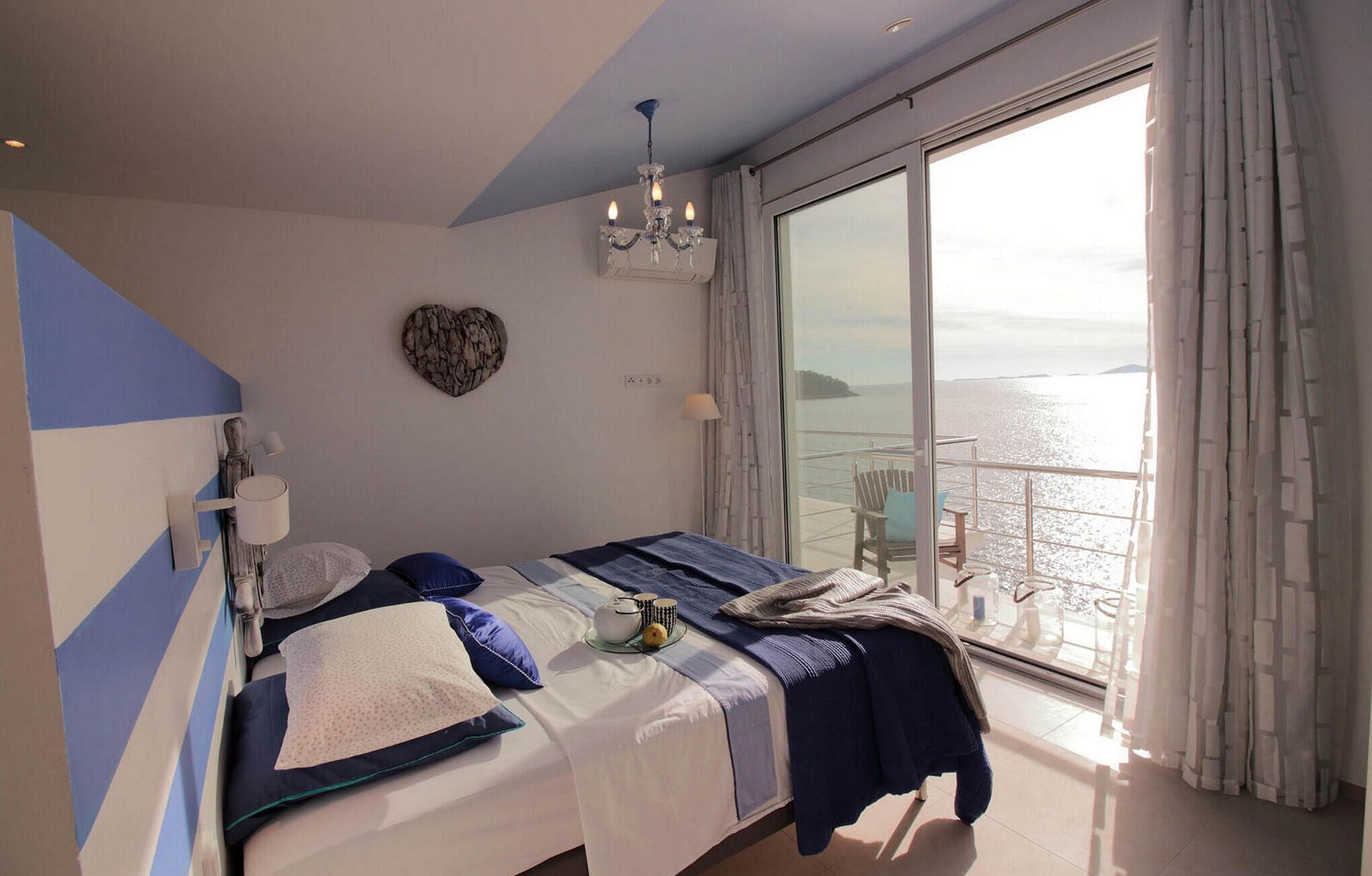 Une chambre à coucher avec un lit, un lustre, un décor mural en forme de cœur et une vue sur la mer grâce à de grandes fenêtres.