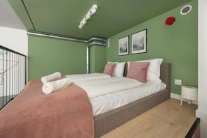 A bedroom with a bed, pillows, a blanket, a small bedside table, a wall-mounted light fixture, and a window with a view.