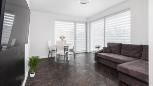 Una sala de estar moderna con una televisión de pantalla plana, un sofá gris, una mesa de comedor con sillas y ventanas grandes con persianas.