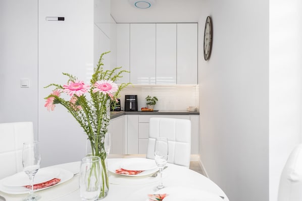 A modern dining area with a white table set for two, a vase with pink flowers, and a kitchen in the background.