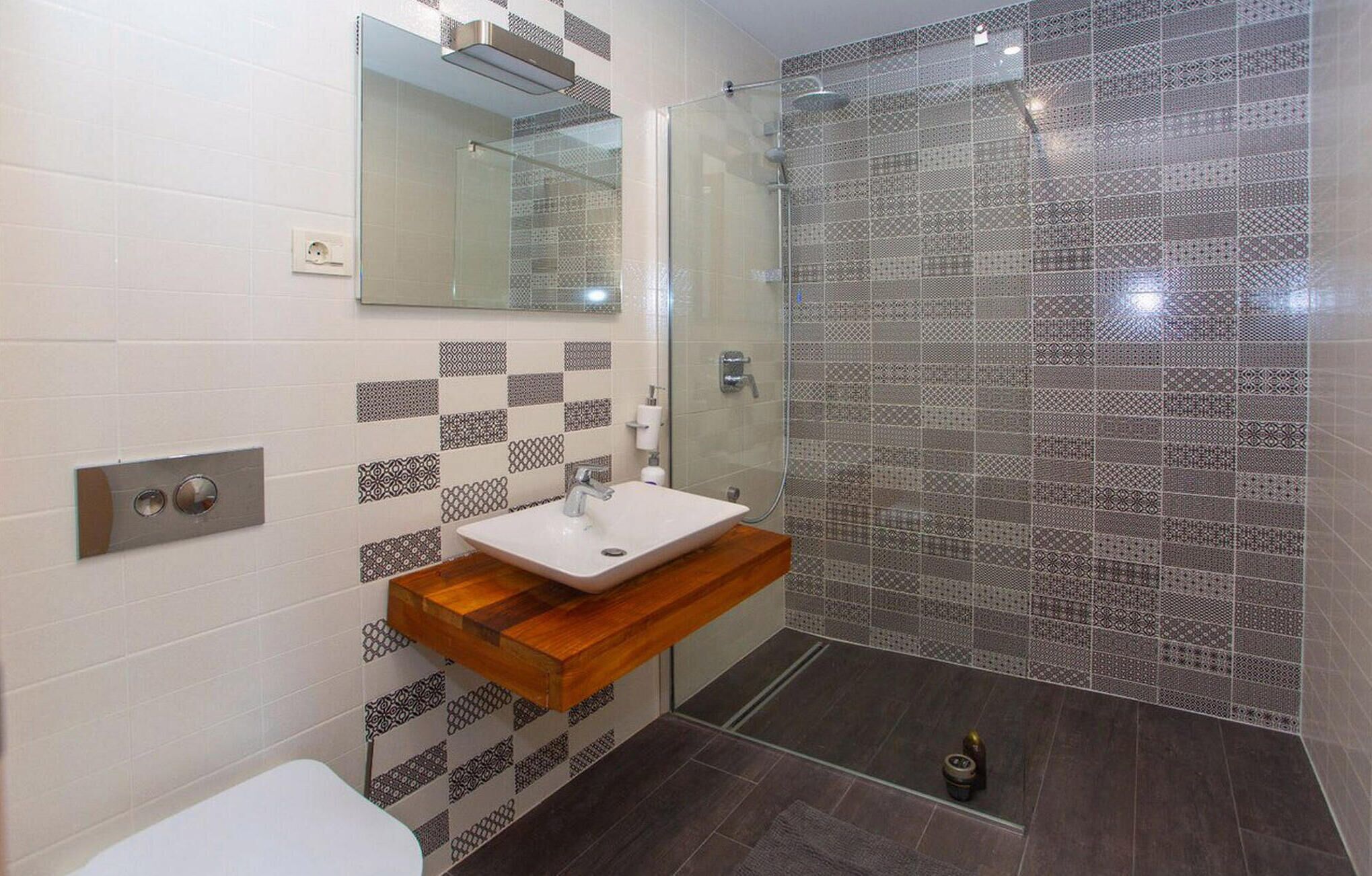 A modern bathroom with a glass shower enclosure, a white sink with a wooden countertop, and patterned tiles on the walls.