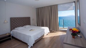 A hotel room with a large bed, a bedside table, a nightstand, a window with a view of the sea, and a small table with a plant and a remote control.