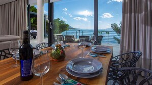 A dining table set with plates, glasses, and a bottle of wine, overlooking a scenic view of the sea and mountains.