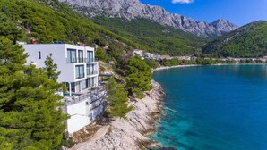 A modern two-story building with a pool, located on a coastal cliff overlooking a clear blue sea and a mountainous backdrop.
