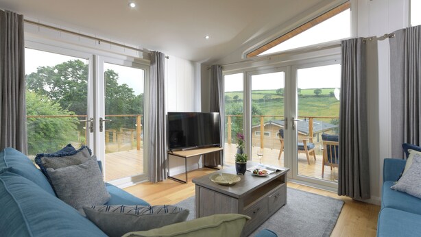 Ein großzügiges Wohnzimmer mit einer großen Couch, einem Couchtisch und einem Fernseher. Das Zimmer verfügt über große Schiebeglasfenster, die auf einen Balkon mit Holzterrasse führen.