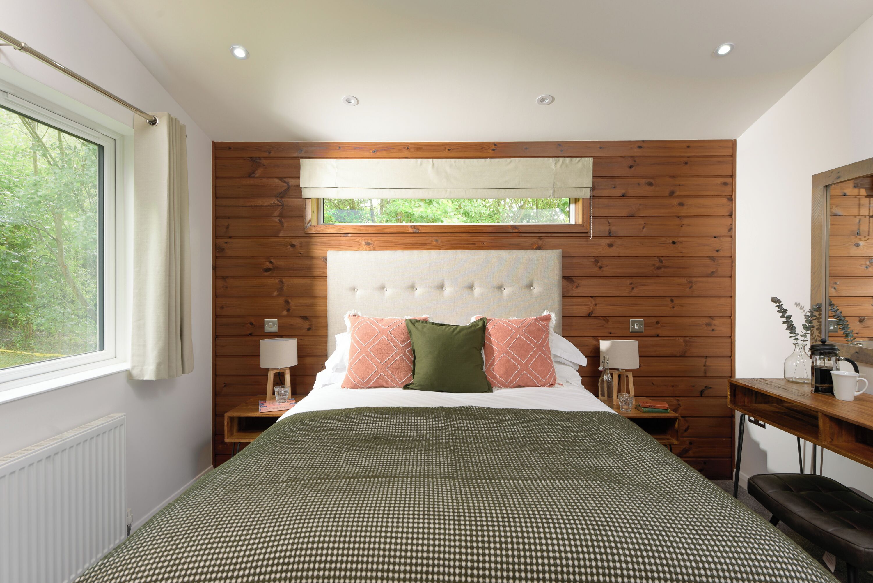 A bedroom with a large bed, wooden panel walls, a window with curtains, two bedside tables with lamps, and a mirror on the wall.