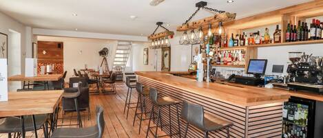 A bar area with wooden flooring, bar stools, a counter with a beverage dispenser, and shelves stocked with bottles.