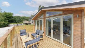 A wooden deck with outdoor furniture, a glass door leading to a covered patio, and a view of a lush green landscape.